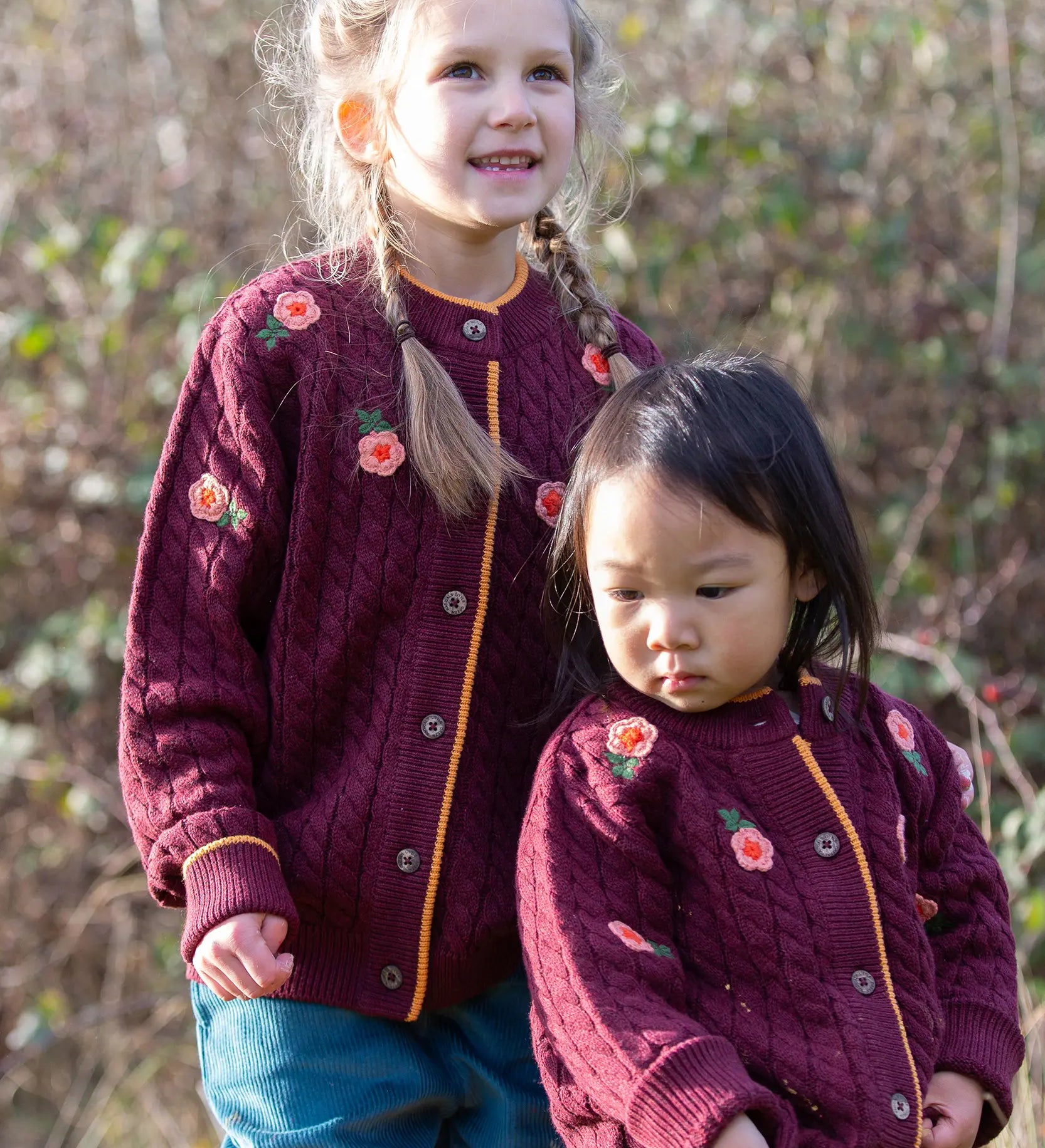 Children wearing the Little Green Radicals organic cotton snuggly knitted cardigan with a berry petal design. Dark purple cardigan with pink flowers details.