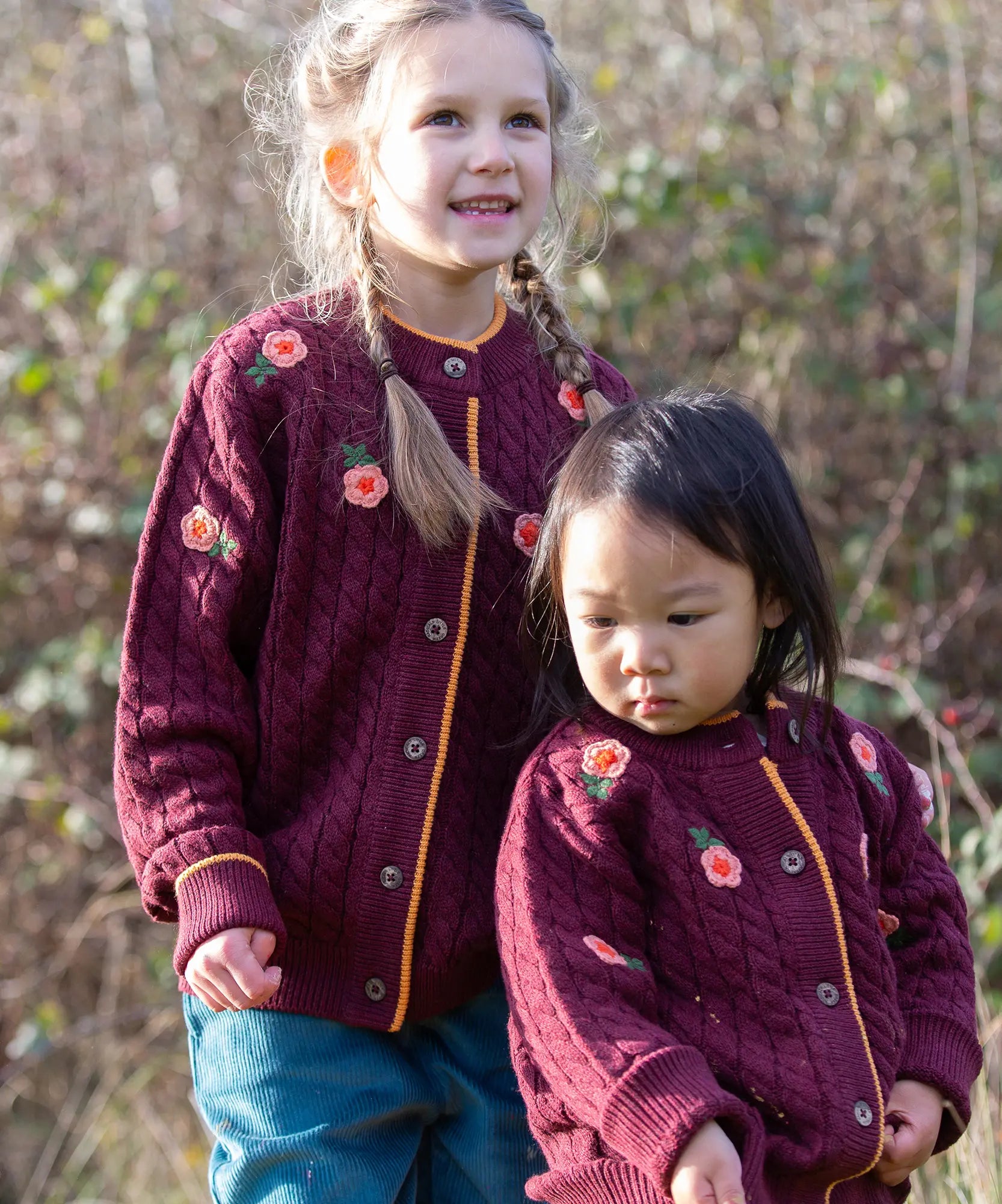 Children wearing the Little Green Radicals organic cotton snuggly knitted cardigan with a berry petal design. Dark purple cardigan with pink flowers details.