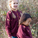 Children wearing the Little Green Radicals organic cotton snuggly knitted cardigan with a berry petal design. Dark purple cardigan with pink flowers details.