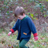 Child wearing the Little Green Radicals organic cotton snuggly knitted cardigan in midnight blue. Navy blue cardigan with red sleeve cuffs, green collar and yellow on the pockets with dark green knee patch joggers.