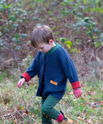 Child wearing the Little Green Radicals organic cotton snuggly knitted cardigan in midnight blue. Navy blue cardigan with red sleeve cuffs, green collar and yellow on the pockets with dark green knee patch joggers.