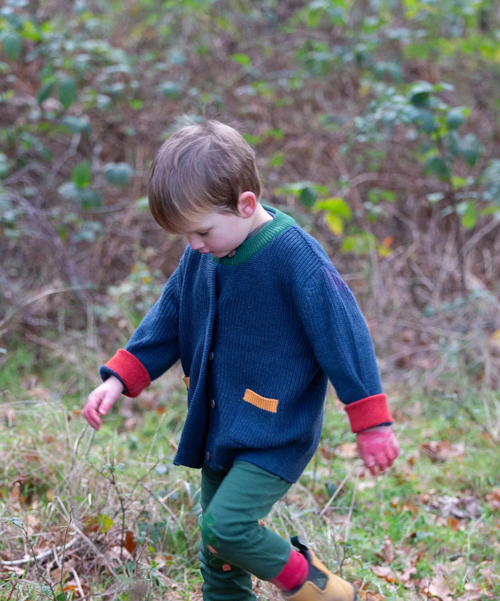 Child wearing the Little Green Radicals organic cotton snuggly knitted cardigan in midnight blue. Navy blue cardigan with red sleeve cuffs, green collar and yellow on the pockets with dark green knee patch joggers.