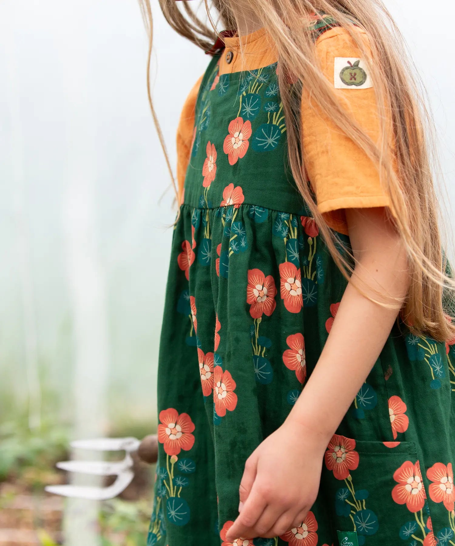 Close up of the side pockets on the Little Green Radicals green orange blooms repeat print reversible pinny kids dress being worn.