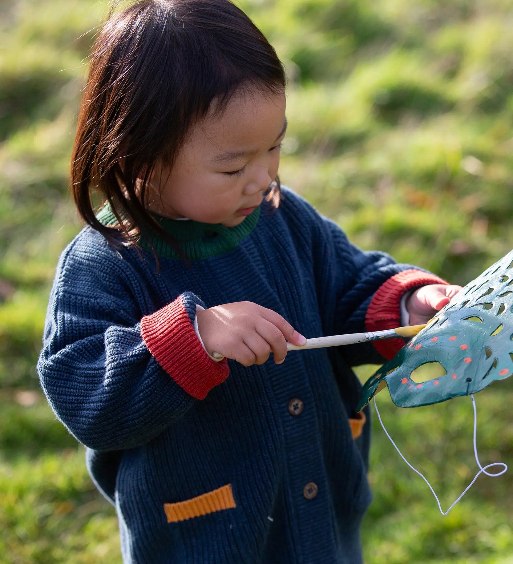 Child wearing the Little Green Radicals organic cotton snuggly knitted cardigan in midnight blue. Navy blue cardigan with red sleeve cuffs, green collar and yellow on the pockets.