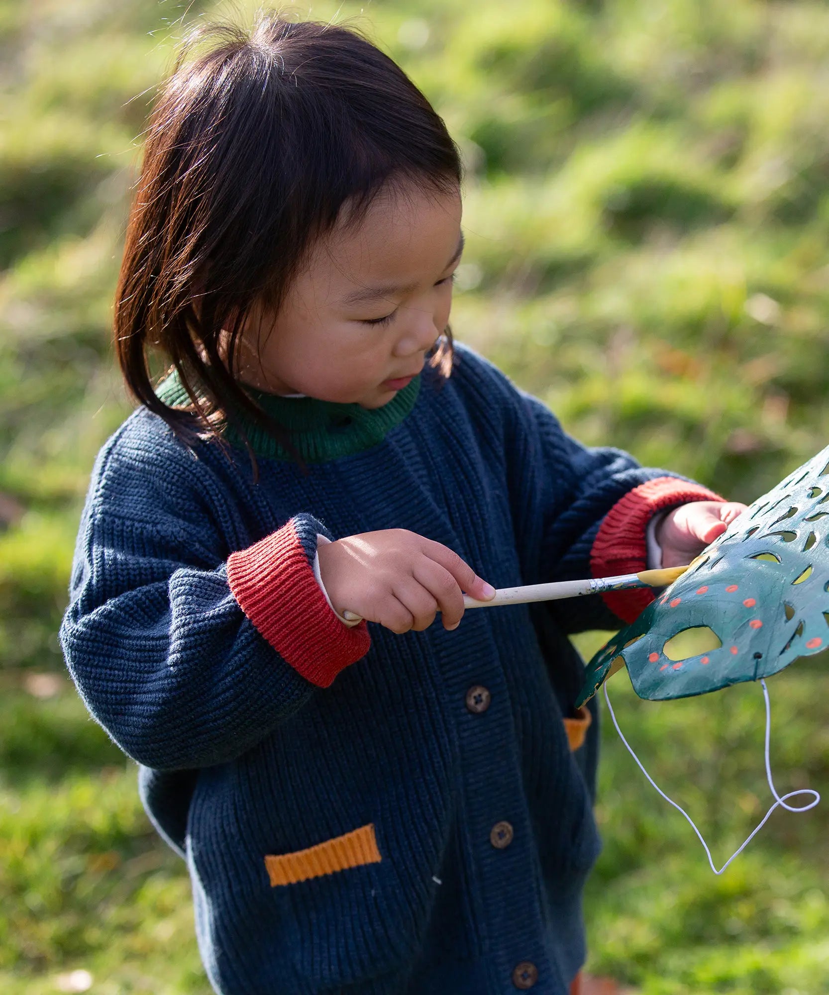 Child wearing the Little Green Radicals organic cotton snuggly knitted cardigan in midnight blue. Navy blue cardigan with red sleeve cuffs, green collar and yellow on the pockets.