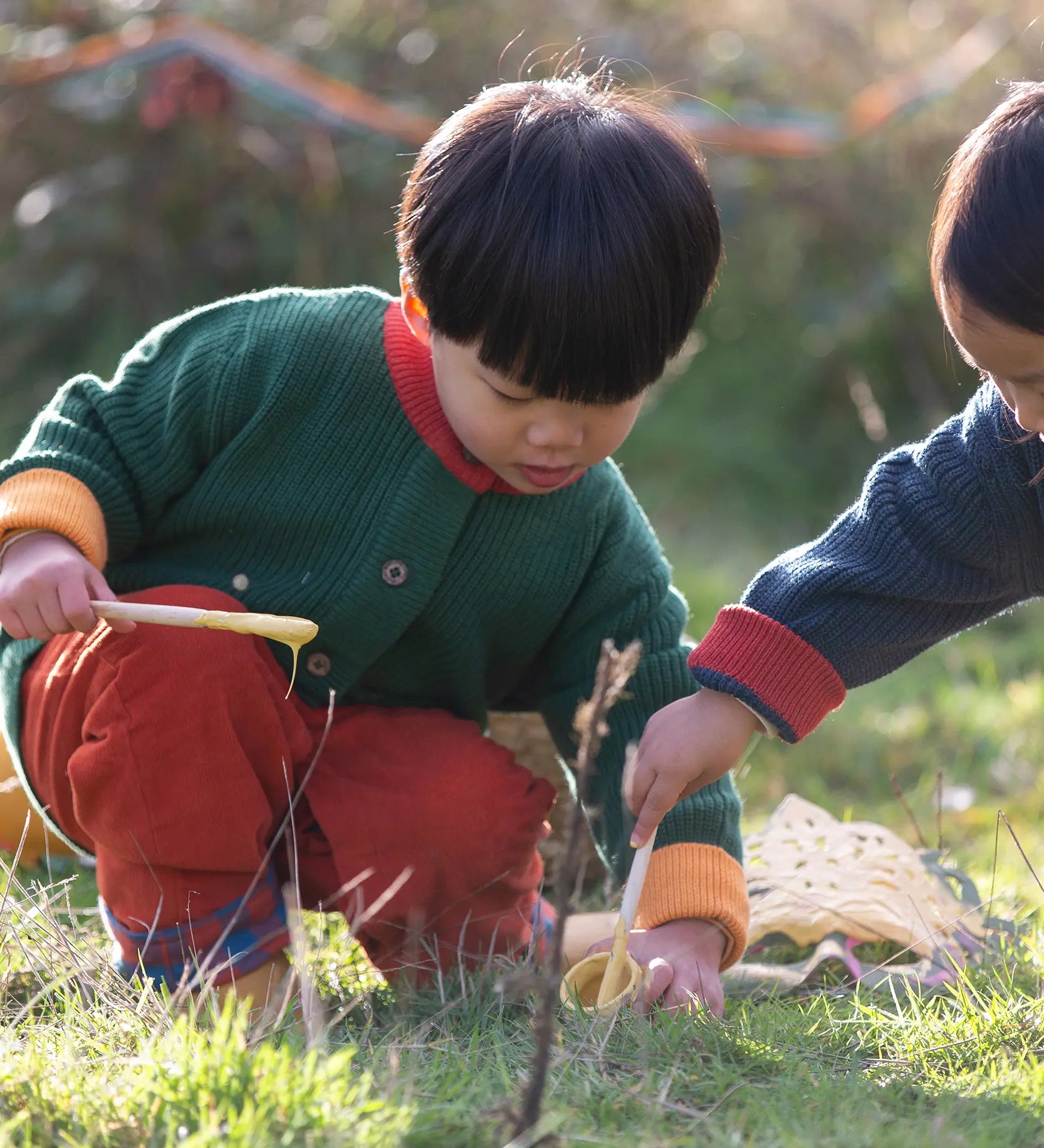 Child wearing the Little Green Radicals organic cotton snuggly knitted cardigan in olive green with orange corduroy trousers kneeling down. 
