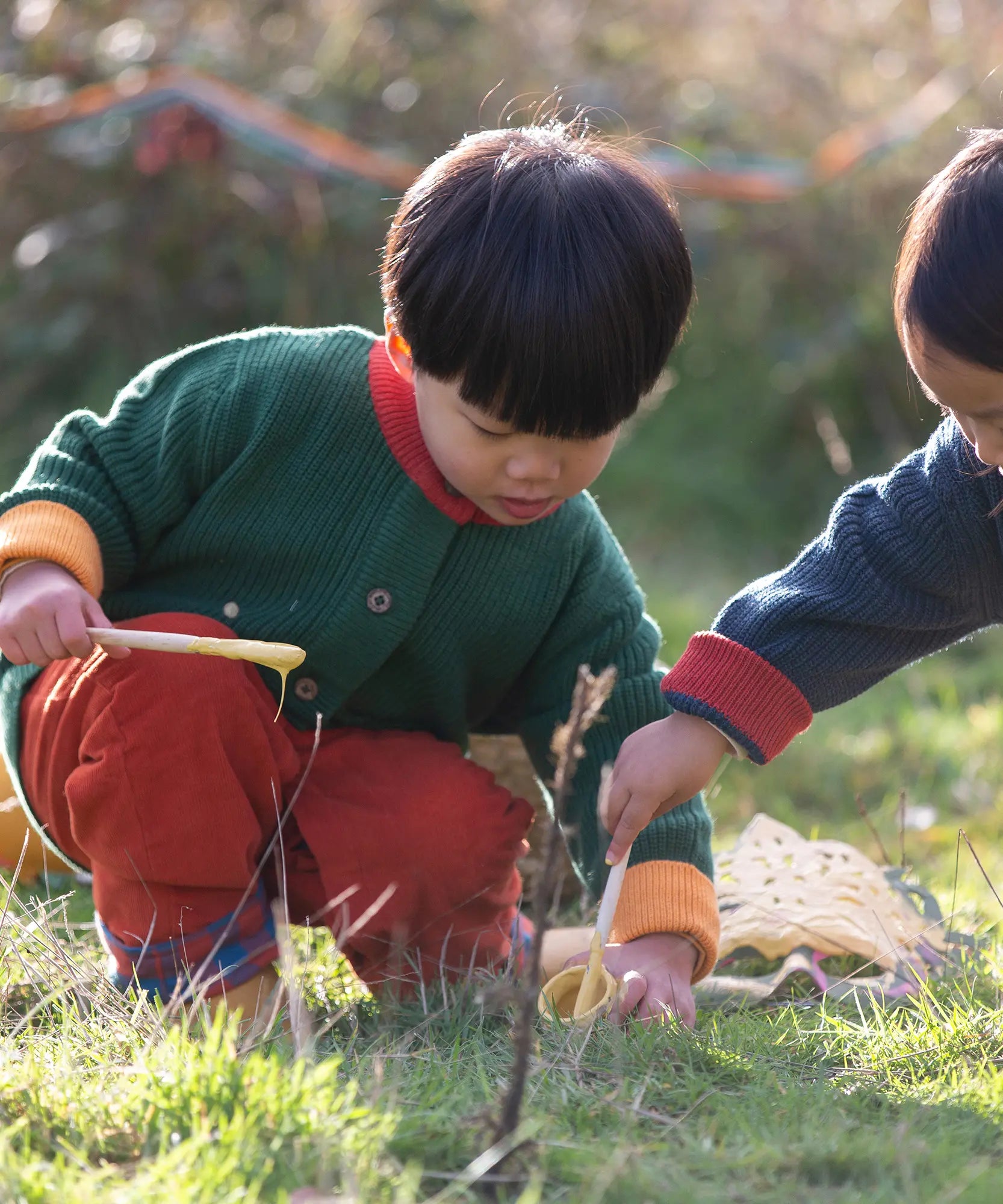 Child wearing the Little Green Radicals organic cotton snuggly knitted cardigan in olive green with orange corduroy trousers kneeling down. 