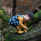 Child wearing Little Green Radicals  blue penguins everyday GOTS organic cotton dungarees, crouching on a large log in a forest. Navy blue background with an all over blue penguin print with yellow gold leg cuffs.