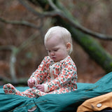 Baby wearing the Little Green Radicals petals GOTS organic cotton zip up babygrow sitting on a green blanket outdoors. Natural coloured cotton with an all over pink flowers print.