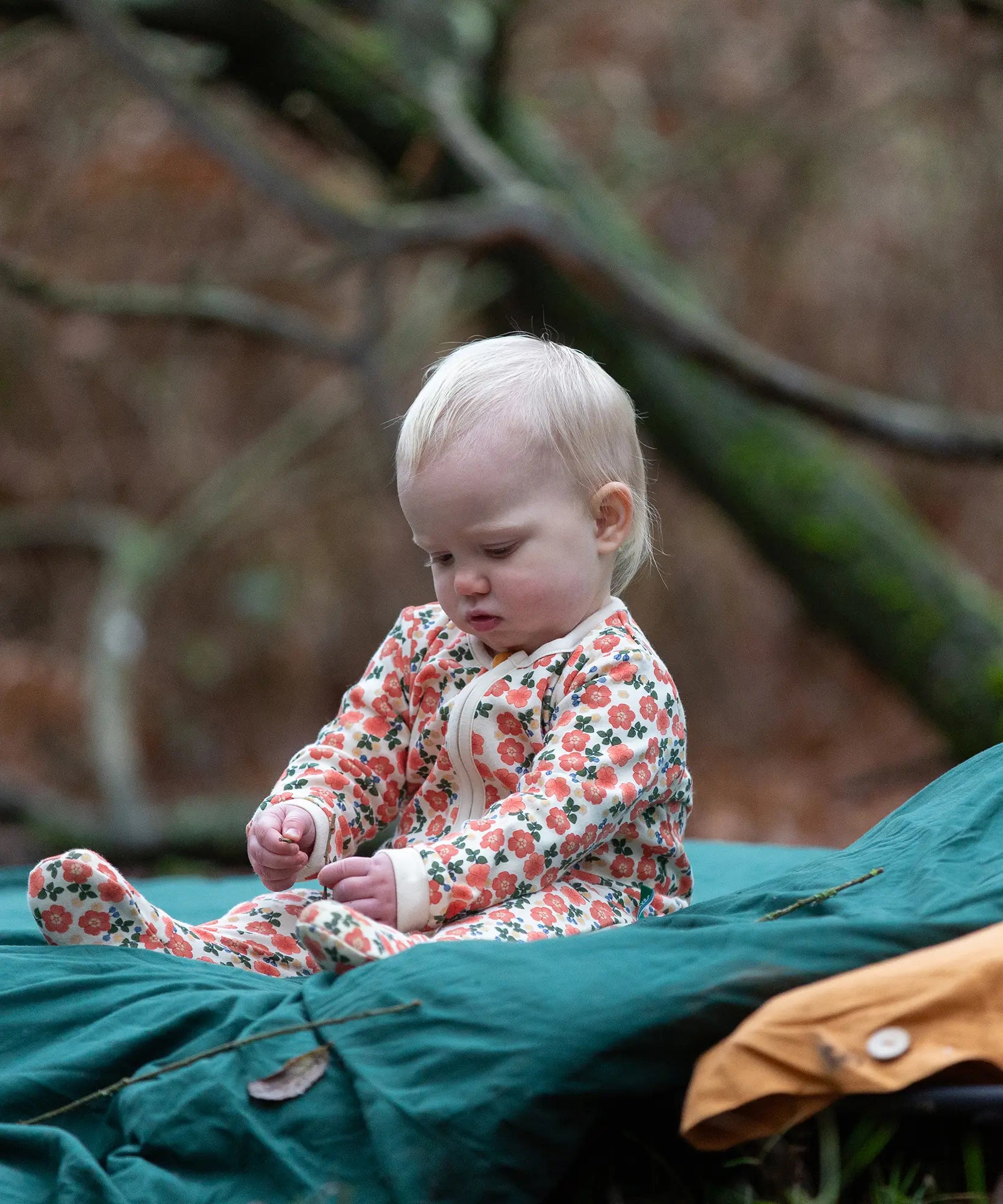 Baby wearing the Little Green Radicals petals GOTS organic cotton zip up babygrow sitting on a green blanket outdoors. Natural coloured cotton with an all over pink flowers print.