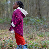 Side of a child wearing a Little Green Radicals pink flowers purple organic cotton sherpa toasty coat with red trousers.
