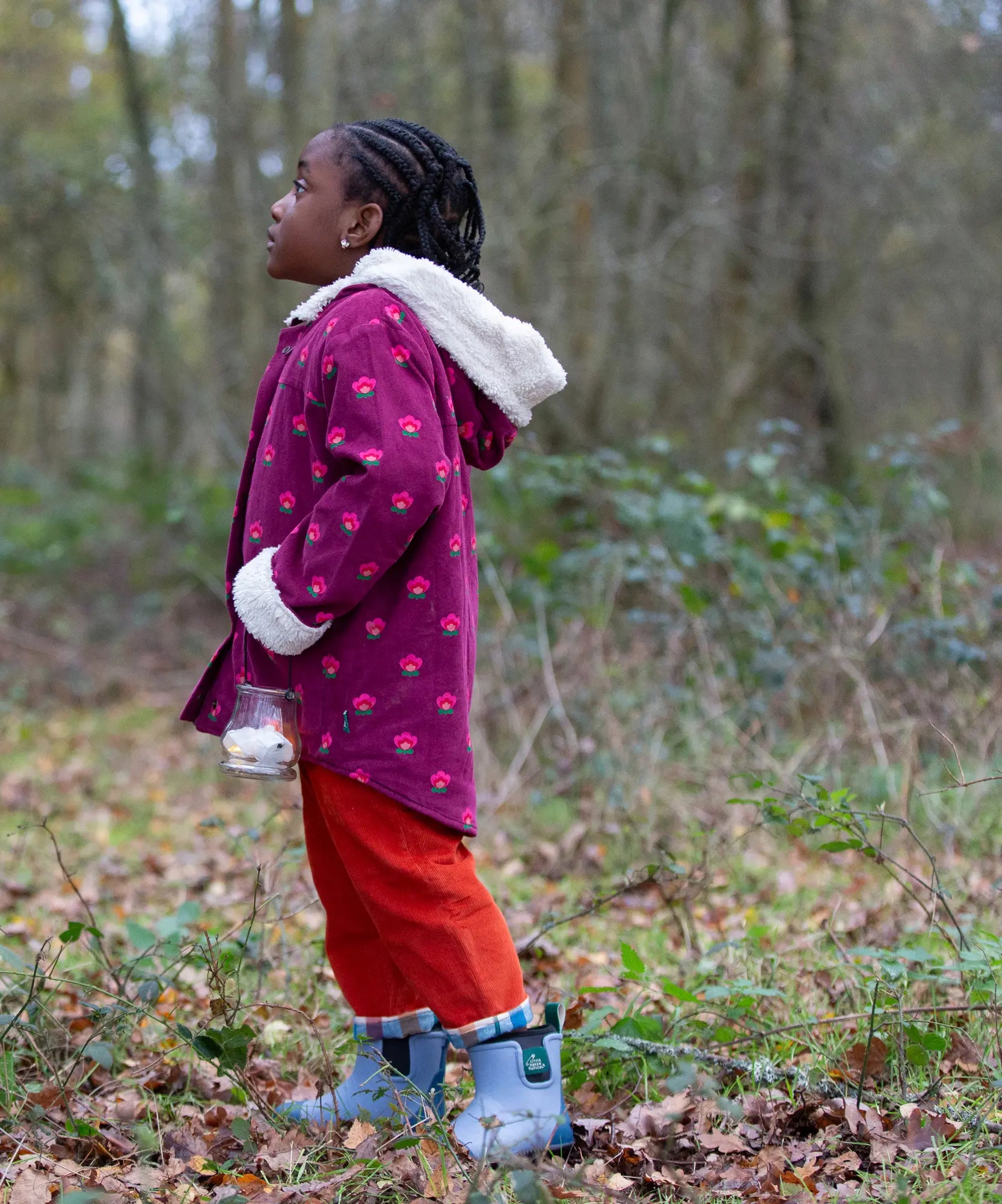 Side of a child wearing a Little Green Radicals pink flowers purple organic cotton sherpa toasty coat with red trousers.