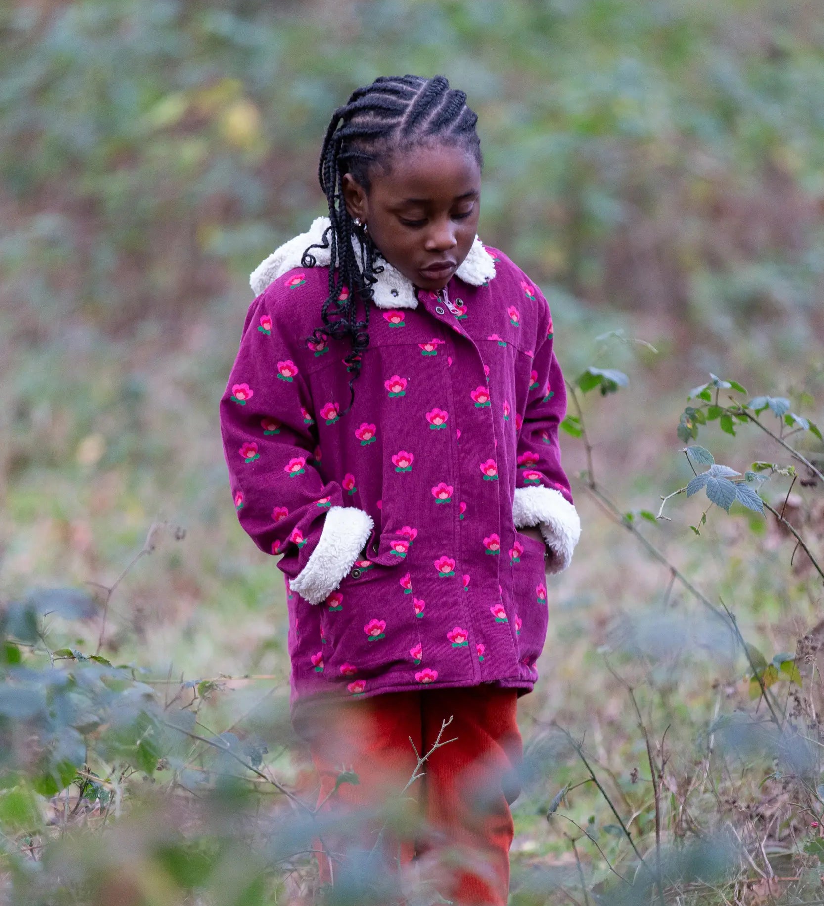 Child wearing a Little Green Radicals pink flowers purple organic cotton sherpa toasty coat with red trousers.