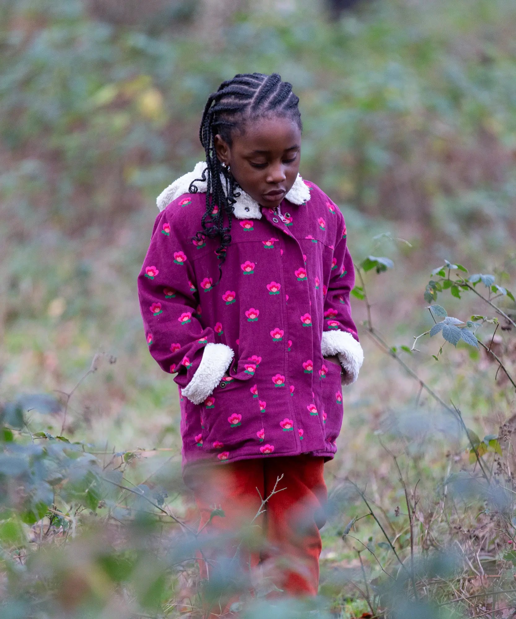 Child wearing a Little Green Radicals pink flowers purple organic cotton sherpa toasty coat with red trousers.