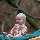 Baby wearing the Little Green Radicals petals GOTS organic cotton zip up babygrow sitting on a green blanket outdoors. Natural coloured cotton with an all over pink flowers print.