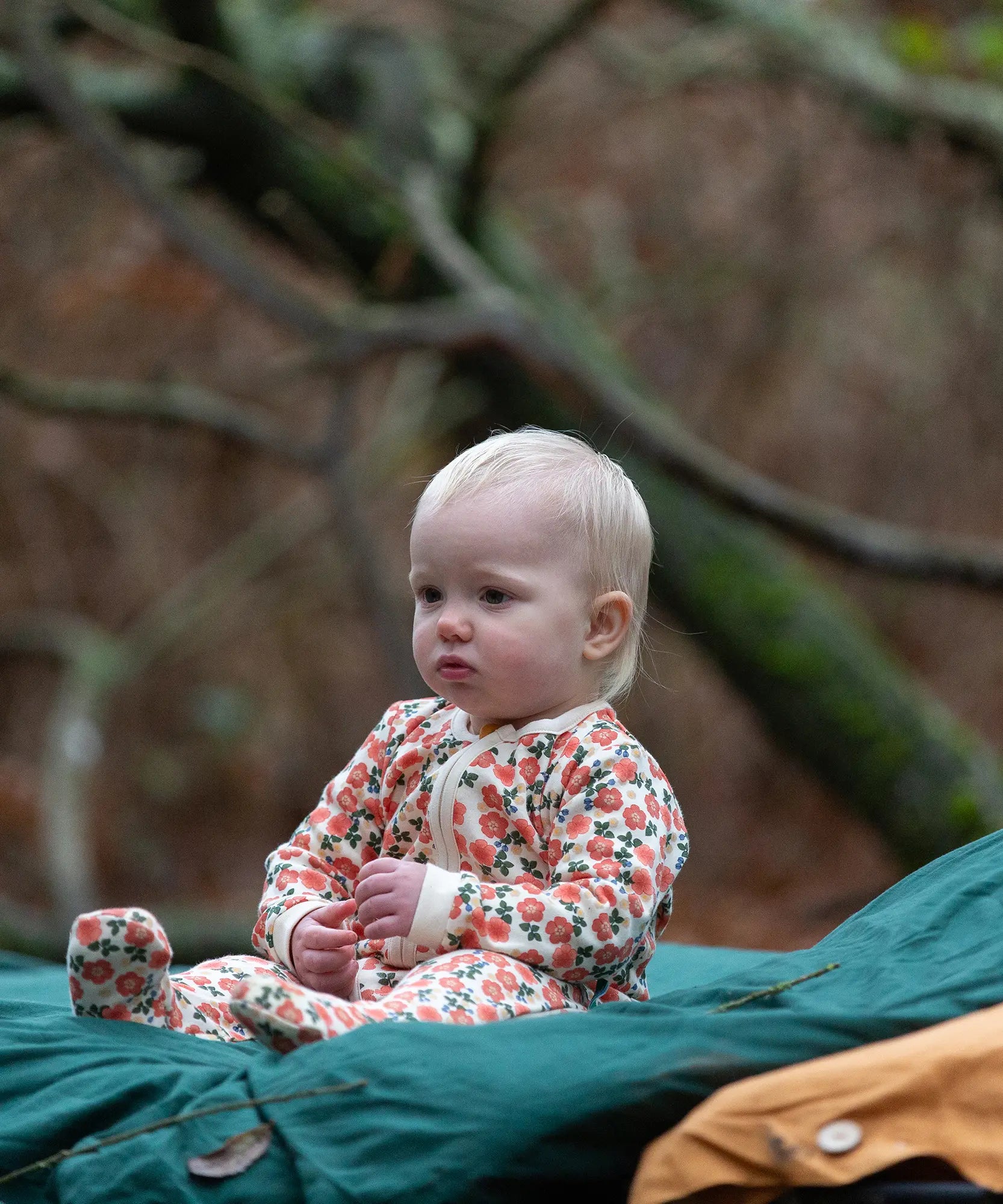 Baby wearing the Little Green Radicals petals GOTS organic cotton zip up babygrow sitting on a green blanket outdoors. Natural coloured cotton with an all over pink flowers print.