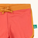 Close up of the Little Green Radicals pink sunset recycled swim shorts with yellow gold waist and binding. A small green logo tag can be seen on the side of the leg. 
