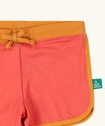 Close up of the Little Green Radicals pink sunset recycled swim shorts with yellow gold waist and binding. A small green logo tag can be seen on the side of the leg. 