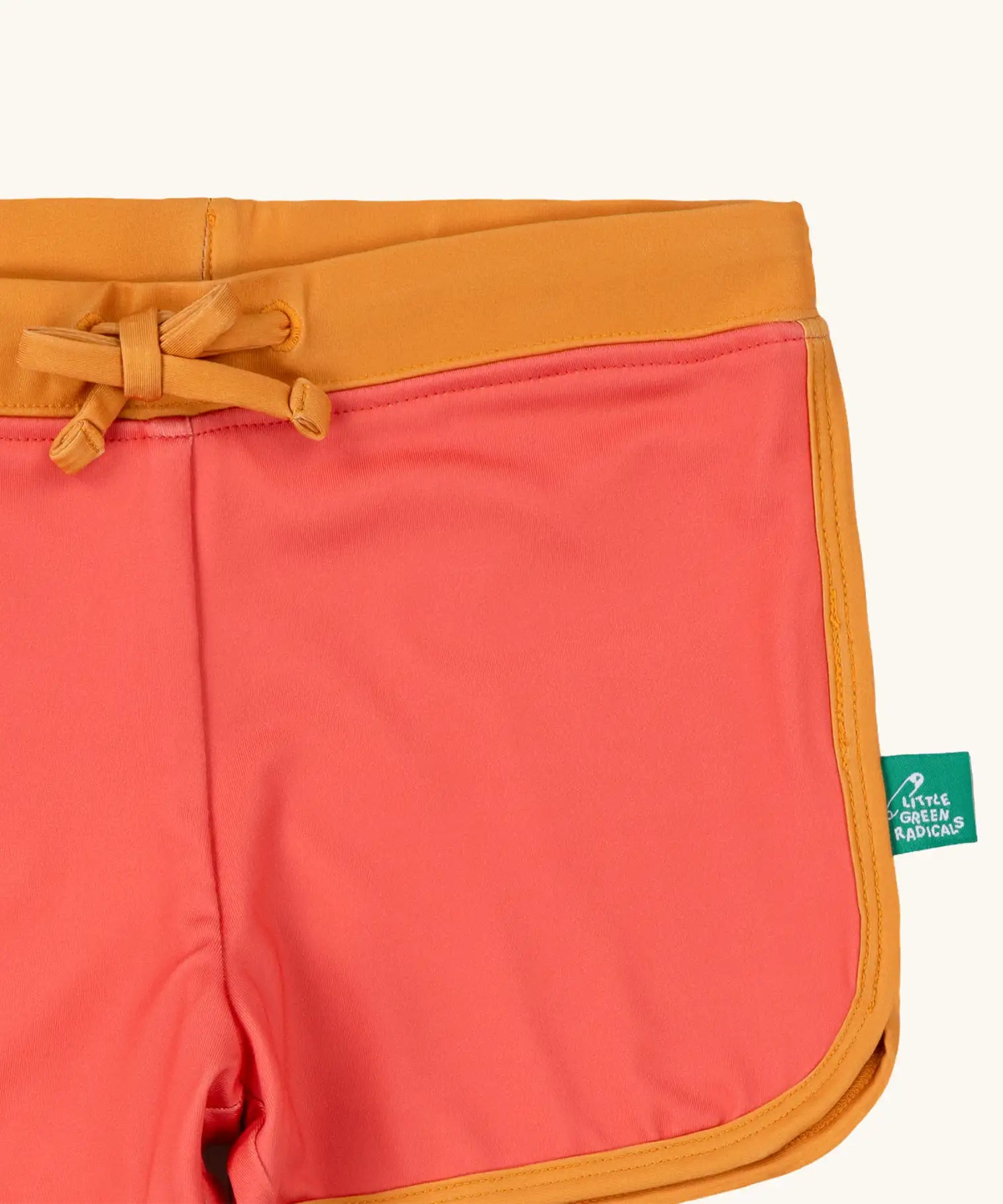 Close up of the Little Green Radicals pink sunset recycled swim shorts with yellow gold waist and binding. A small green logo tag can be seen on the side of the leg. 