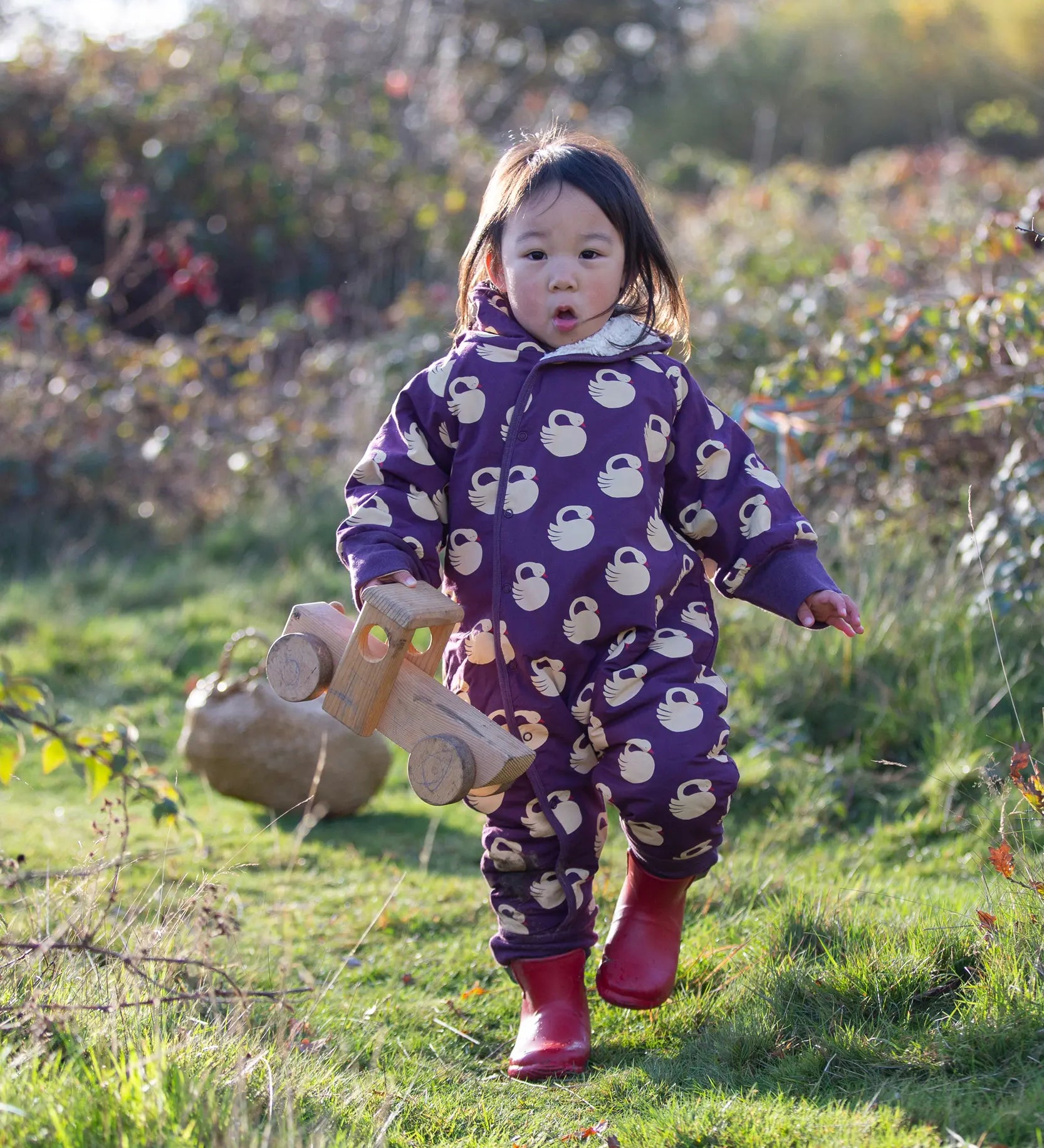 Child wearing the Little Green Radicals purple sleeping swans print sherpa lined snowsuit with red wellington boots.