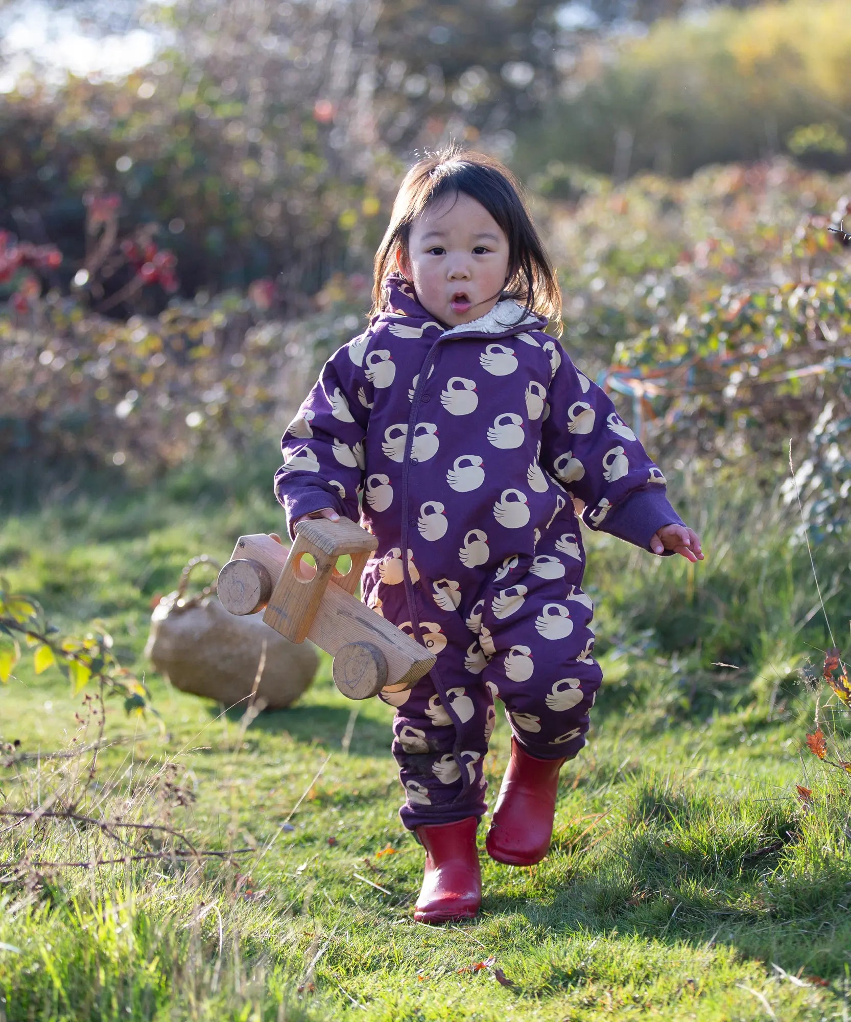 Child wearing the Little Green Radicals purple sleeping swans print sherpa lined snowsuit with red wellington boots.