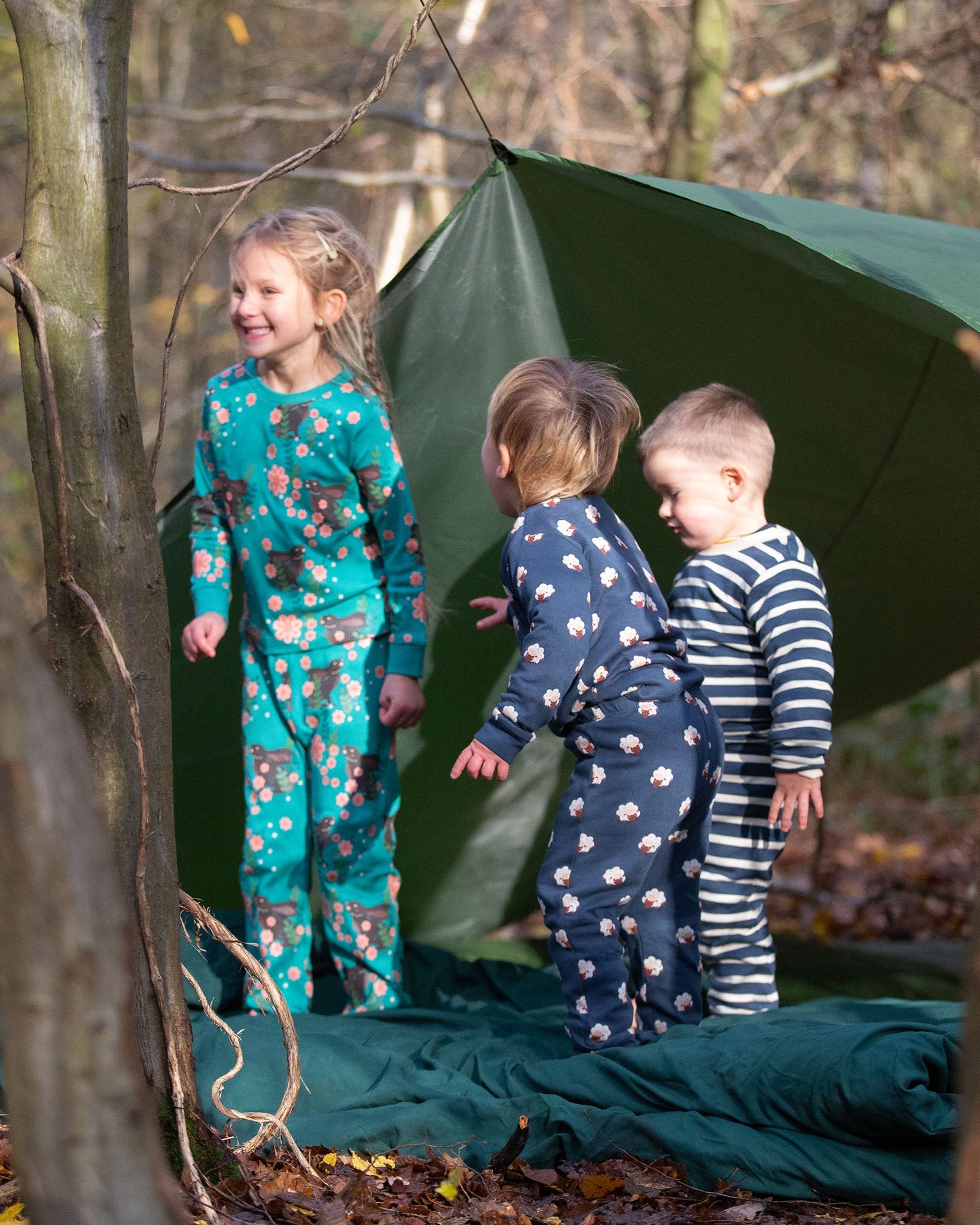 Children standing outside a tent in the forest. They are all wearing Little Green Radicals organic cotton pyjamas. One is wearing a pair with the Winter Rabbit on them, one in wearing navy and white striped design and the third is wearing the sleeping sheep print. Little green Radicals have a wide range of organic cotton, Fairtrade children's clothing an outerwear available here at Babipur.