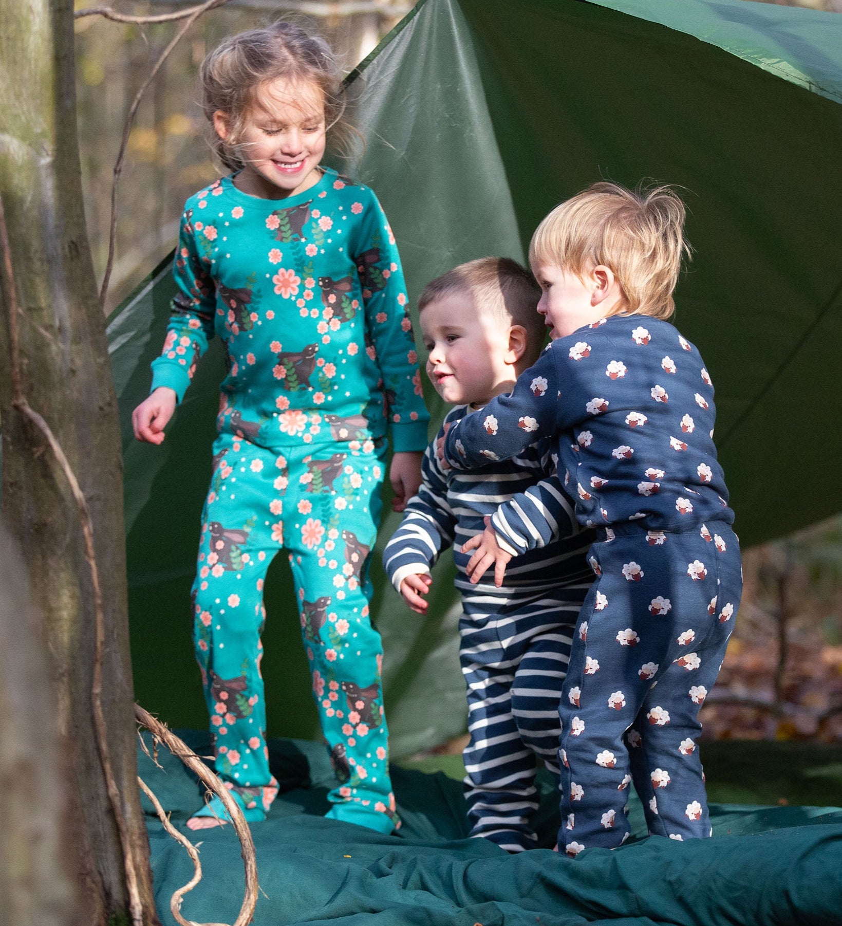 Children standing outside a tent in the forest. They are all wearing Little Green Radicals organic cotton Fairtrade pyjamas. One is wearing a pair with the Winter Rabbit on them, one in wearing navy and white striped design and the third is wearing the sleeping sheep print. Little green Radicals have a wide range of organic cotton, Fairtrade children's clothing an outerwear available here at Babipur.