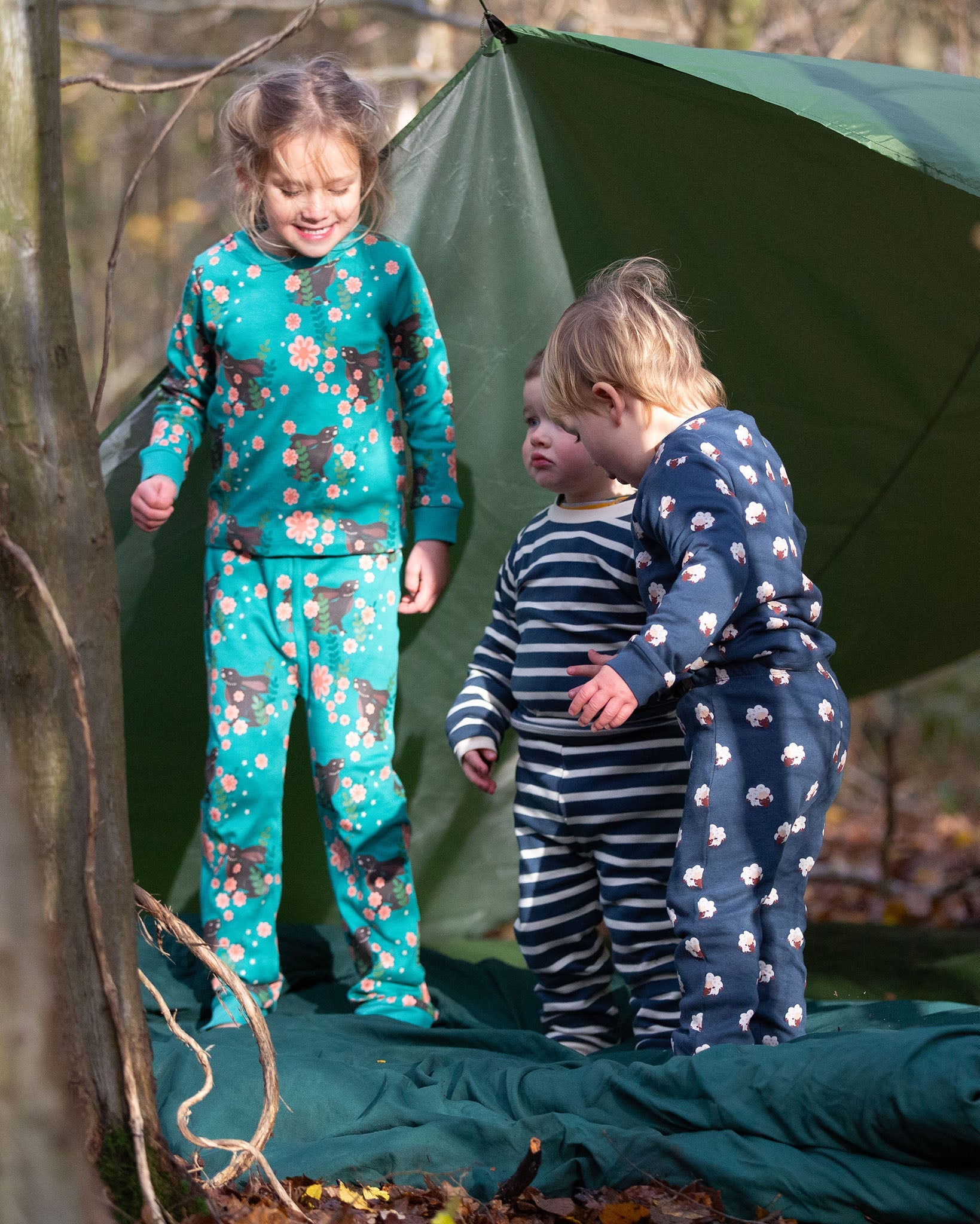 Children standing outside a tent in the forest. They are all wearing Little Green Radicals organic ccotton pyjamas. One is wearing a pair with the Winter Rabbit on them, one in wearing navy and white striped design and the third is wearing the sleeping sheep print. Little green Radicals have a wide range of organic cotton, Fairtrade children's clothing an outerwear available here at Babipur.