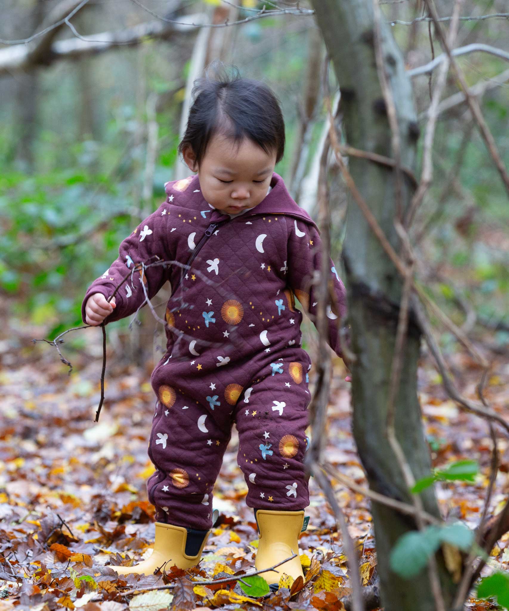 A child wearing the Little Green Radicals Organic Cotton Flying Birds Quilted Snug Suit. A maroon brown coloured suit with an all over print featuring flying birds, stars, moon and  sun. This snug suit is a part of a wide range of organic cotton clothing for children available here at Babipur. 
