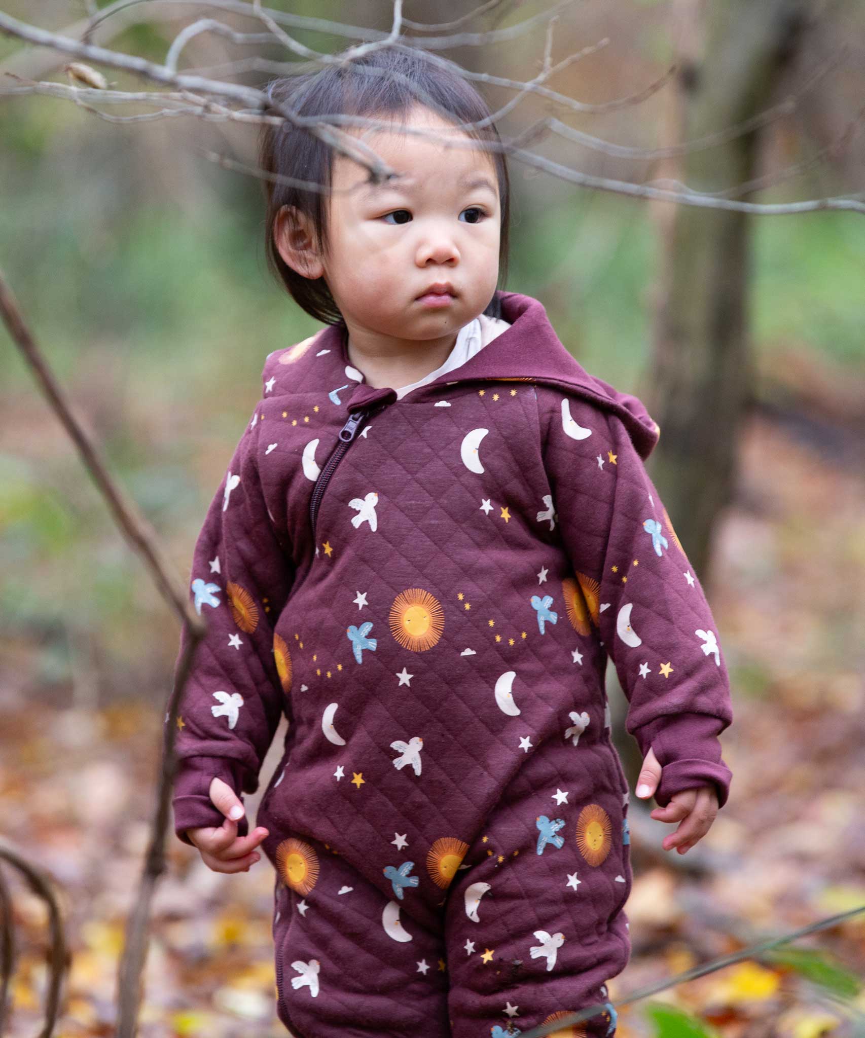 A child wearing the Little Green Radicals Organic Cotton Flying Birds Quilted Snug Suit. A maroon brown coloured suit with an all over print featuring flying birds, stars, moon and  sun. This snug suit is a part of a wide range of organic cotton clothing for children available here at Babipur. 