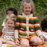 Child wearing the Little Green Radicals kids spring rainbow striped knitted button up organic cotton cardigan with yellow gold shorts. Another child is sitting besides her in a dress.,