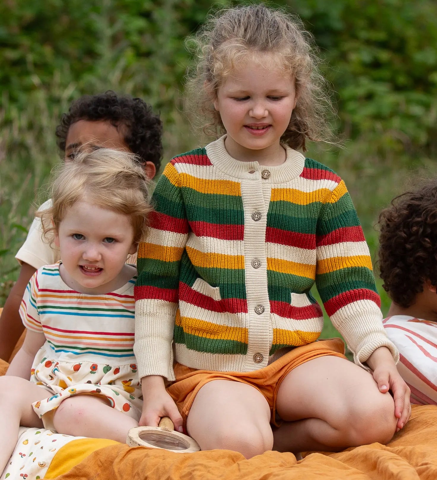 Child wearing the Little Green Radicals kids spring rainbow striped knitted button up organic cotton cardigan with yellow gold shorts. Another child is sitting besides her in a dress.,