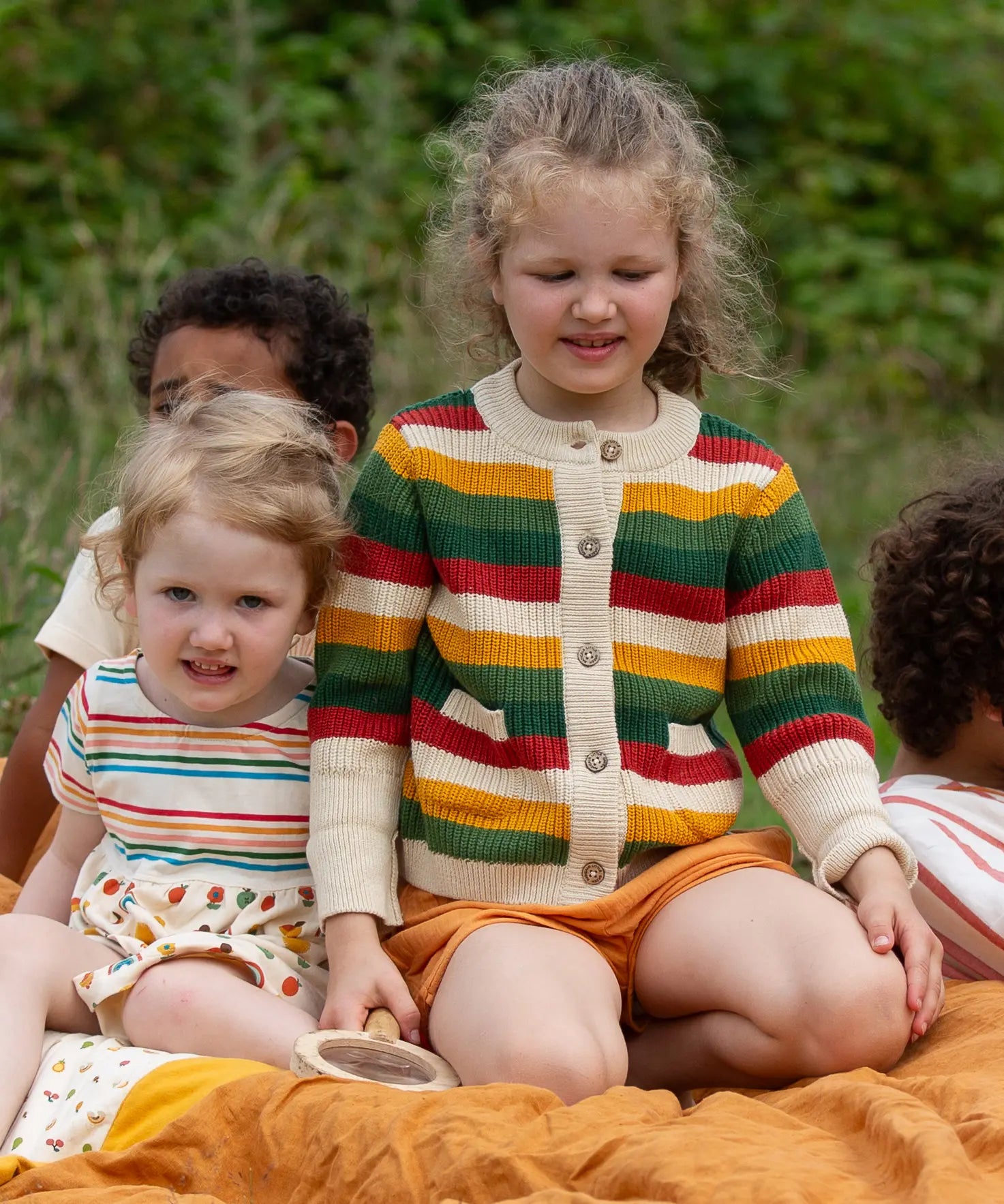 Child wearing the Little Green Radicals kids spring rainbow striped knitted button up organic cotton cardigan with yellow gold shorts. Another child is sitting besides her in a dress.,