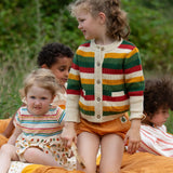 Child wearing the Little Green Radicals kids spring rainbow striped knitted button up organic cotton cardigan with yellow gold shorts. Another child is sitting besides her in a dress.,