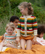 Child wearing the Little Green Radicals kids spring rainbow striped knitted button up organic cotton cardigan with yellow gold shorts. Another child is sitting besides her in a dress.,
