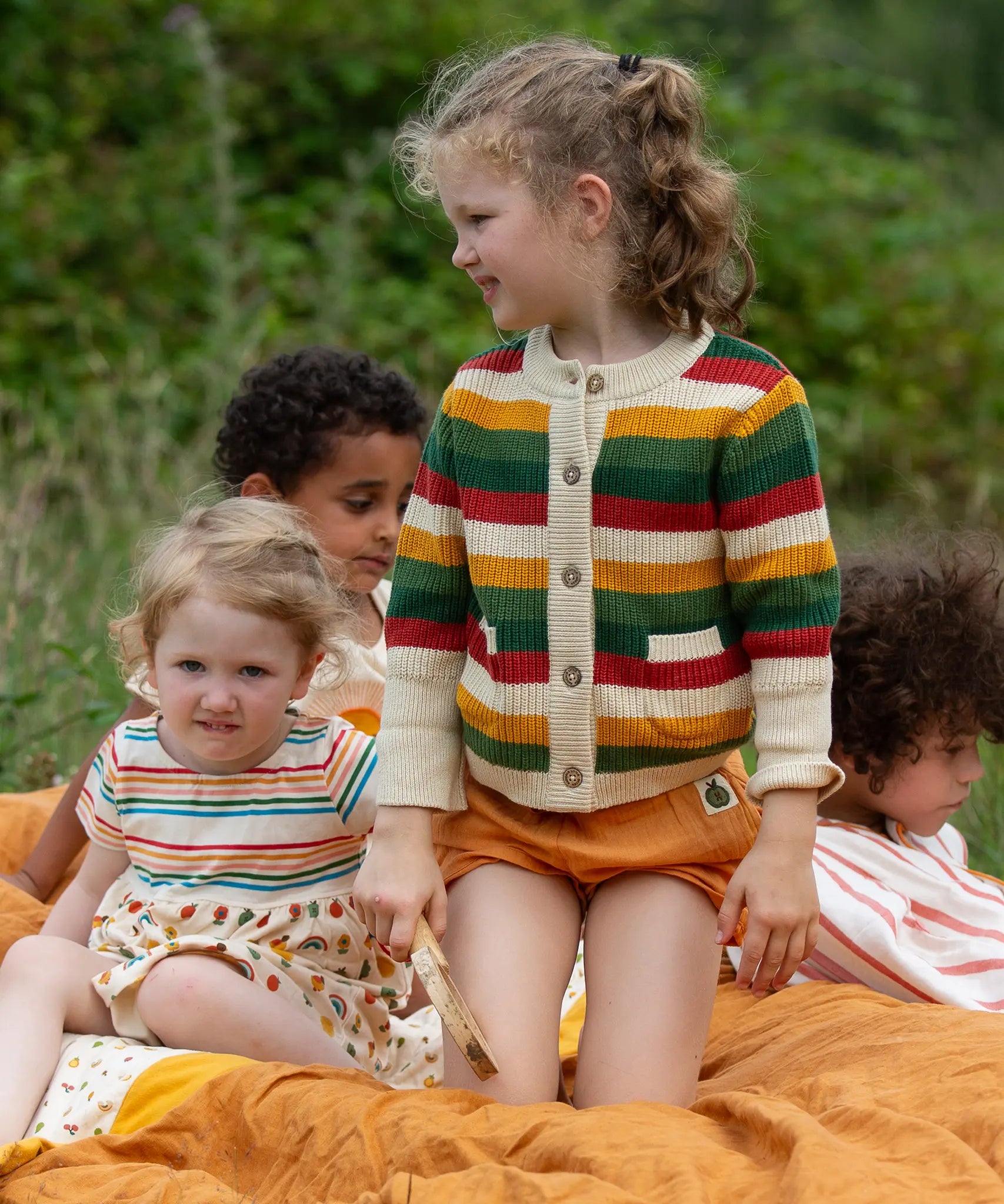 Child wearing the Little Green Radicals kids spring rainbow striped knitted button up organic cotton cardigan with yellow gold shorts. Another child is sitting besides her in a dress.,