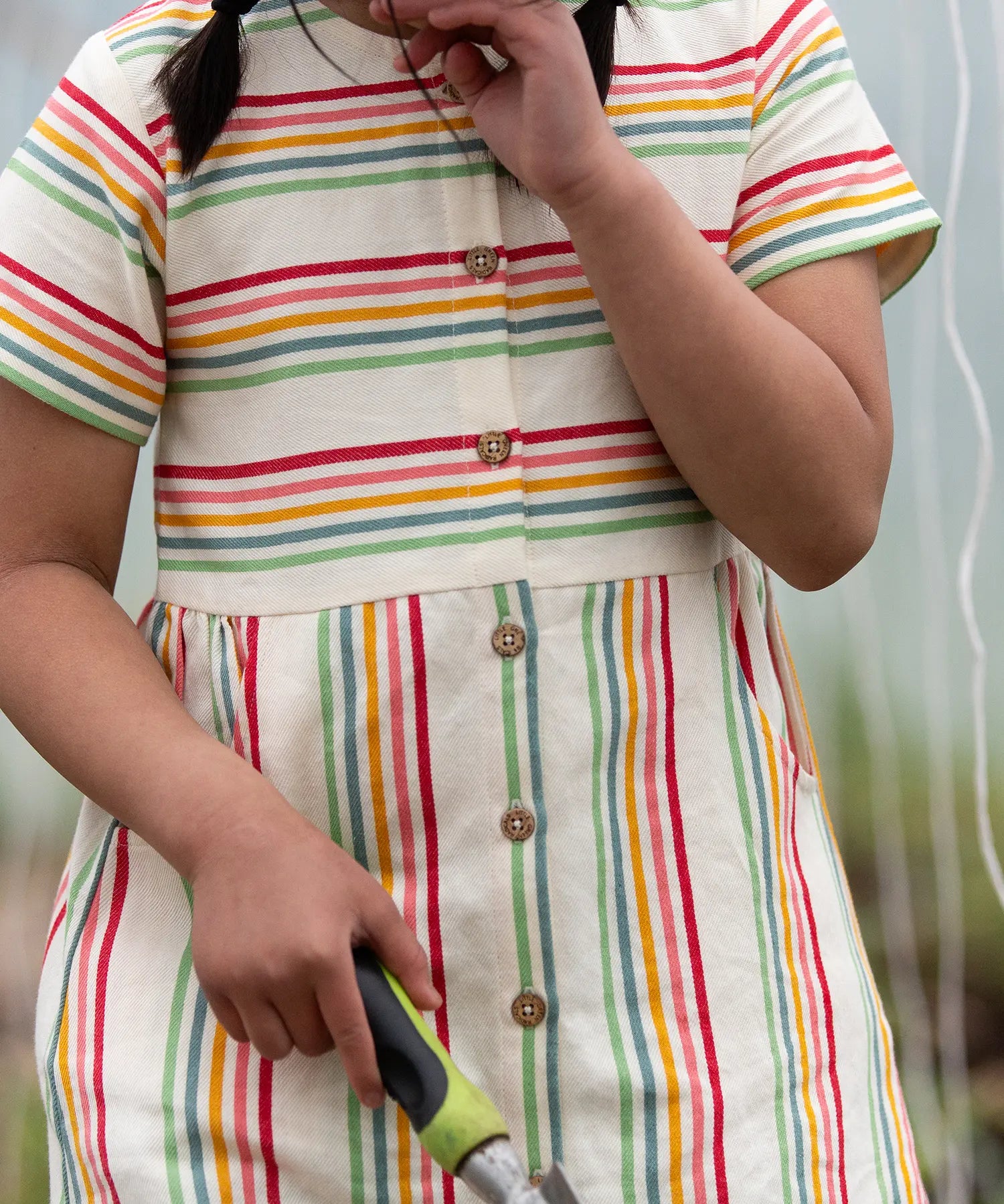 A close up of the button up front on the Little Green Radicals children's cream rainbow striped short sleeve dress being worn by a child.