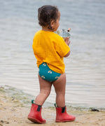 A young child wearing a Little Green Radicals Teal Rainbows UPF 50+ Recycled Reusable Swim Nappy with a yellow gold coloured t-shirt. The child is facing away from the camera.