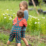 Back of a child wearing the Little Green Radicals kids red apple print blue dungaree shorts with burnt ochre red muslin Henley t-short underneath.