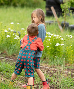 Back of a child wearing the Little Green Radicals kids red apple print blue dungaree shorts with burnt ochre red muslin Henley t-short underneath.