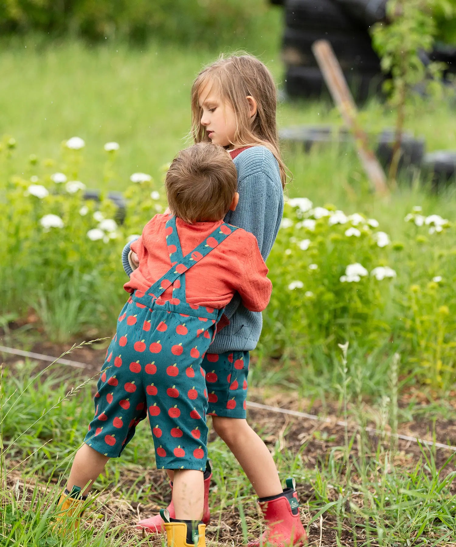 Back of a child wearing the Little Green Radicals kids red apple print blue dungaree shorts with burnt ochre red muslin Henley t-short underneath.