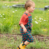 Child wearing the Little Green Radicals kids red apple print blue dungaree shorts with burnt ochre red muslin Henley t-short underneath with yellow gold wellington boots.