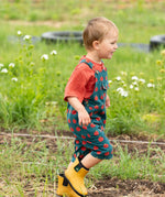 Child wearing the Little Green Radicals kids red apple print blue dungaree shorts with burnt ochre red muslin Henley t-short underneath with yellow gold wellington boots.