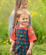 Child wearing the Little Green Radicals kids red apple print blue dungaree shorts with burnt ochre red muslin Henley t-short underneath.