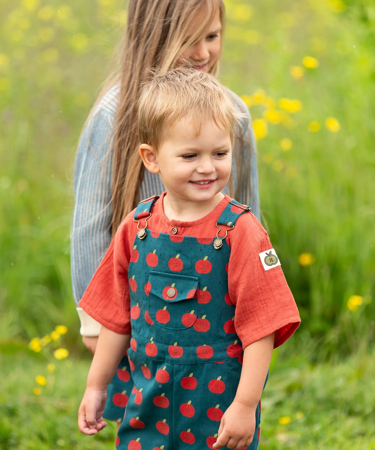 Child wearing the Little Green Radicals kids red apple print blue dungaree shorts with burnt ochre red muslin Henley t-short underneath.