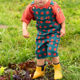 Child wearing the Little Green Radicals kids red apple print blue dungaree shorts with burnt ochre red muslin Henley t-shirt underneath and yellow wellington boots.