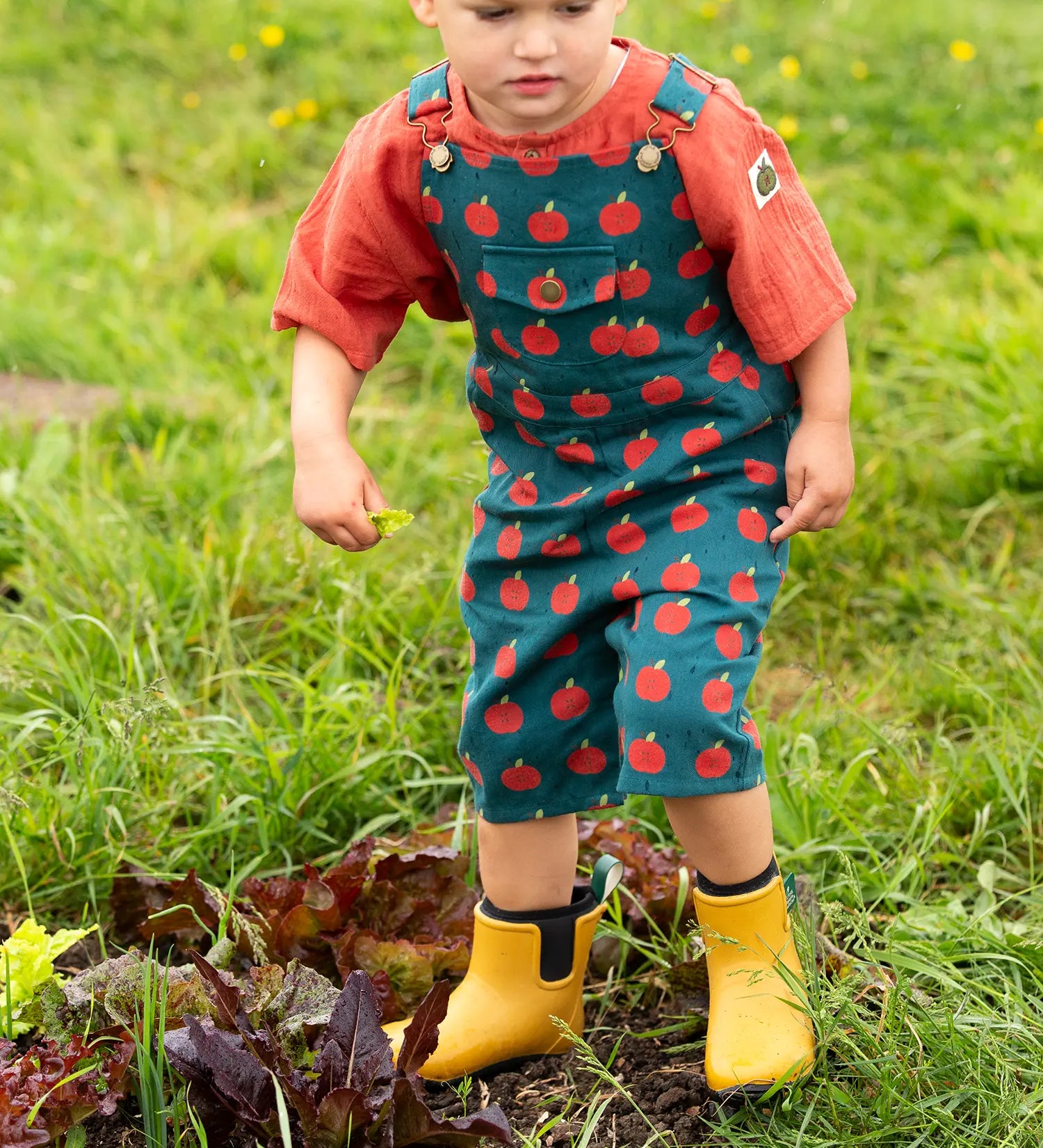 Child wearing the Little Green Radicals kids red apple print blue dungaree shorts with burnt ochre red muslin Henley t-shirt underneath and yellow wellington boots.