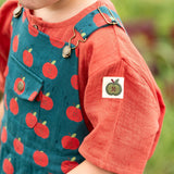 Child wearing the Little Green Radicals kids red apple print blue dungaree shorts with burnt ochre red muslin Henley t-short underneath.
