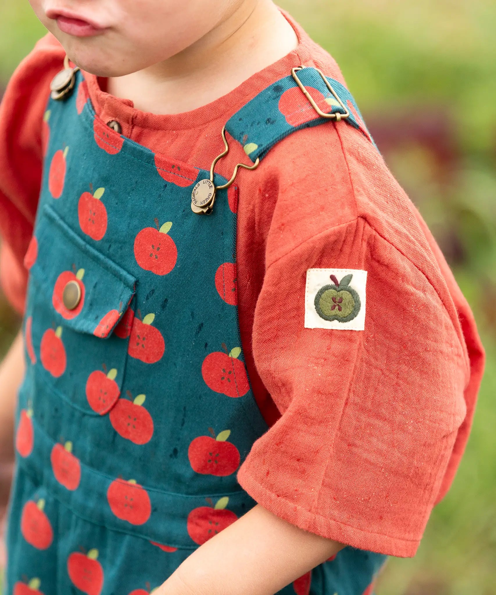 Little Green Radicals kids red apple print blue dungaree shorts with burnt ochre red muslin Henley t-shirt underneath on a child.