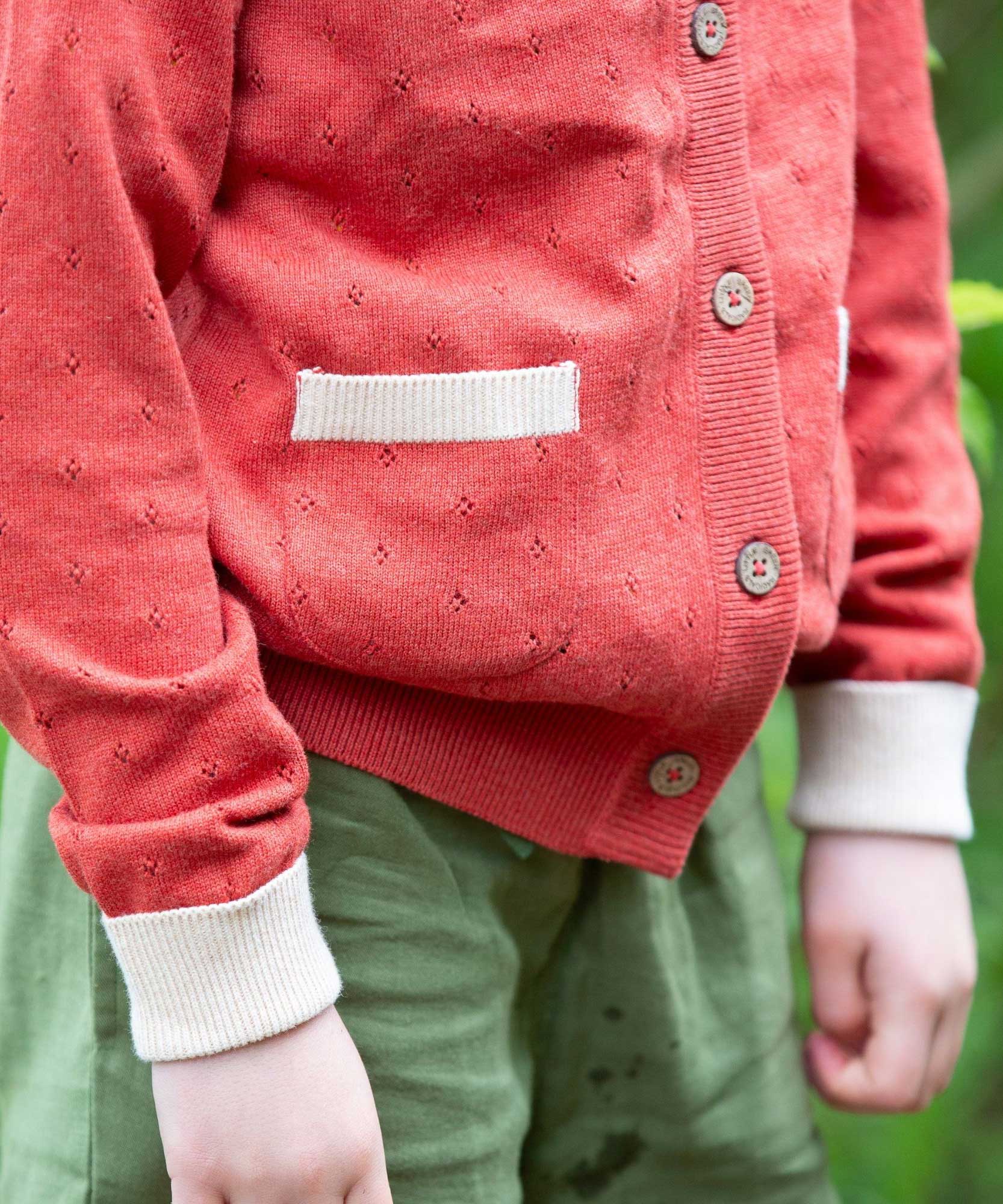 A close up of the pocket on the Little Green Radicals From One To Another Burnt Ochre Pointelle Knitted Cardigan with green coloured shorts. Little green Radicals have a wide range of organic cotton, Fairtrade children's clothing and outerwear available here at Babipur.